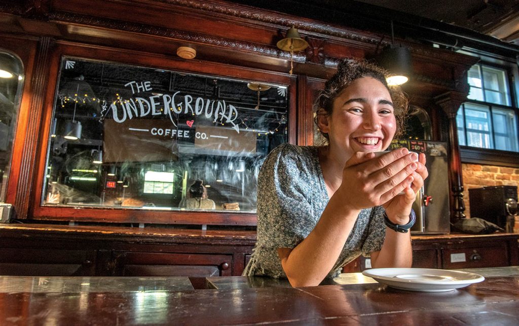 Image of Tevah Gevelber leaning on the bar with a cup of coffee in her hands