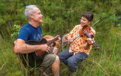 Image of Rory MacLeod and Sandol Astrausky playing the guitar and fiddle in a green field by trees.