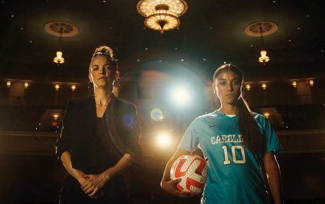 Image of two women standing on a stage with spotlights behind them, one holding a soccer ball.