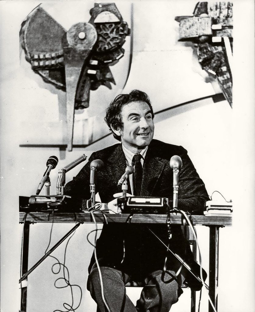 Archival image of Leon Cooper sitting at table with microphones in front of him.
